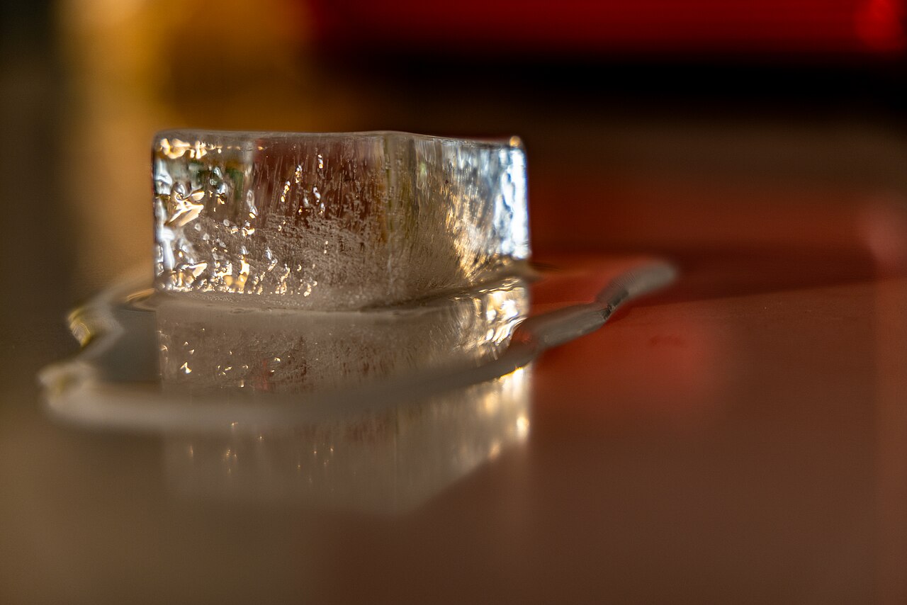 A single ice cube resting in a puddle of meltwater on a reflective surface.